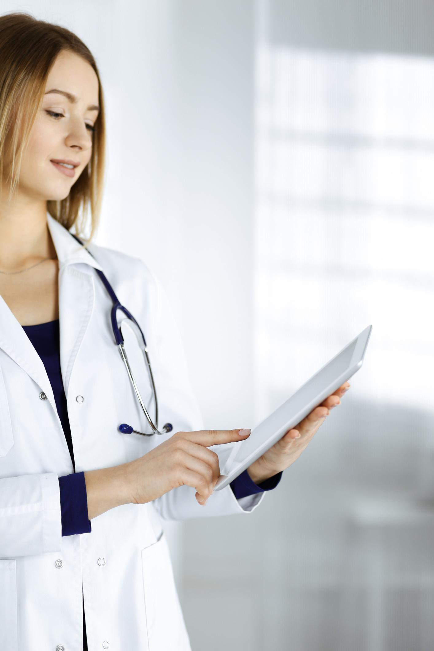 Young woman-doctor is holding a tablet computer in her hands, while standing in a clinic.